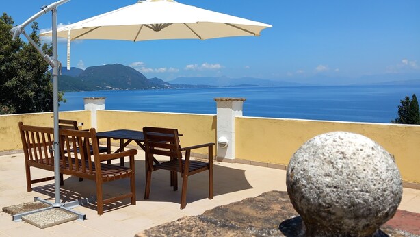 Un espace extérieur aménagé avec une table et des chaises sous un parasol blanc, offrant une vue panoramique sur la mer et les montagnes lointaines.
