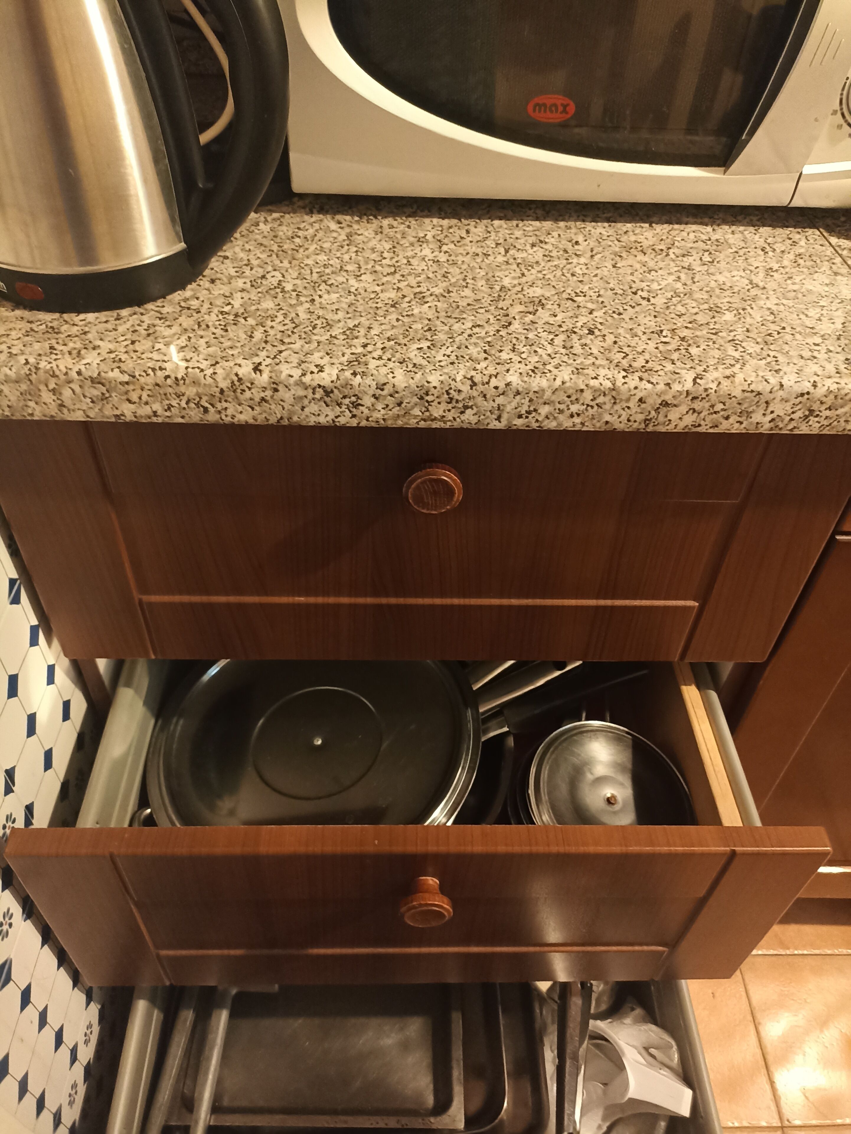 A kitchen with a granite countertop, a microwave, a kettle, and a drawer containing pots and pans.