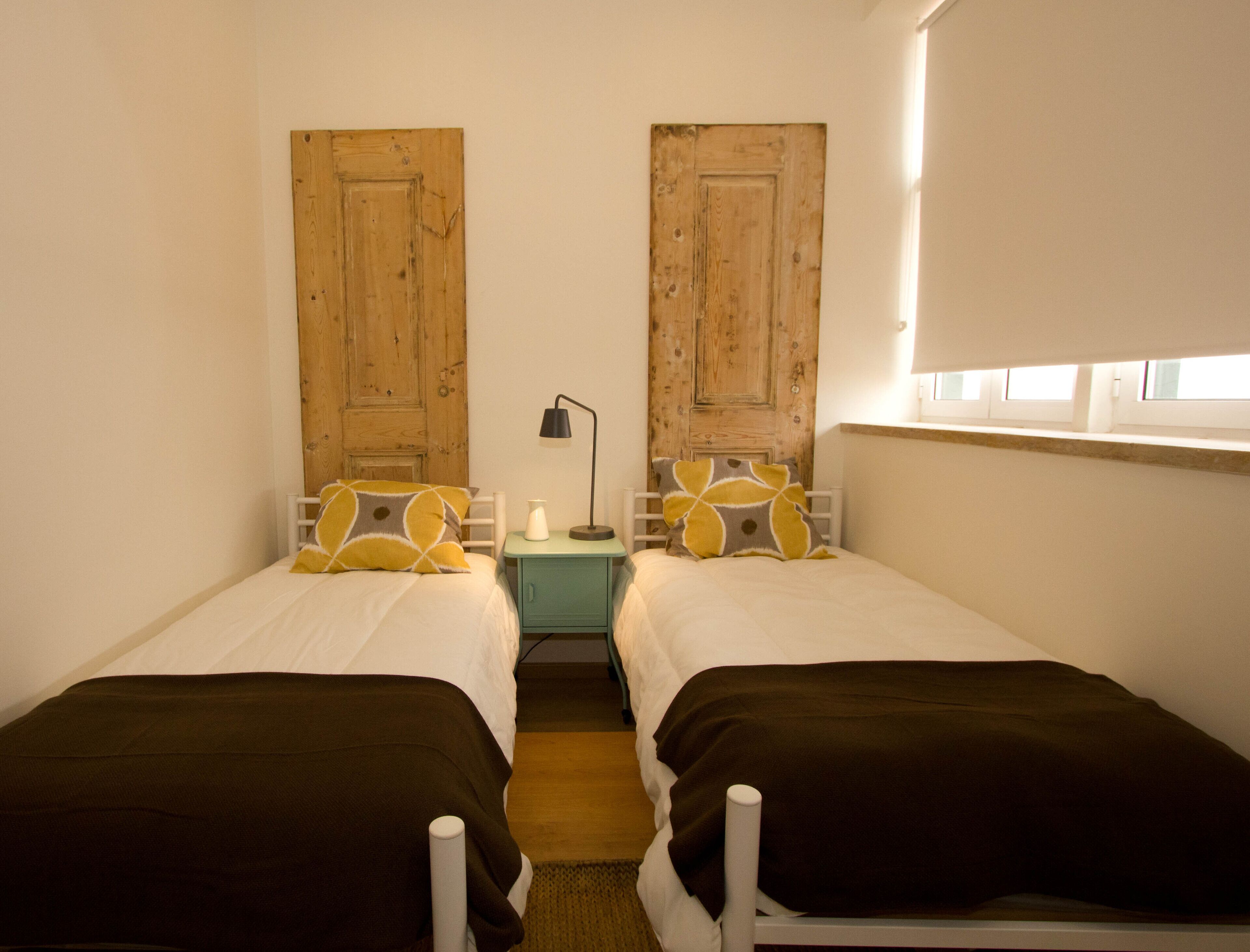 Una habitación pequeña y acogedora con dos camas individuales, una mesita con una lámpara y una ventana con persianas.