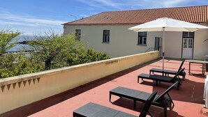 Eine Dachterrasse mit Liegestühlen und einem weißen Sonnenschirm, mit Blick auf ein Gebäude mit einem Ziegeldach und Meerblick.