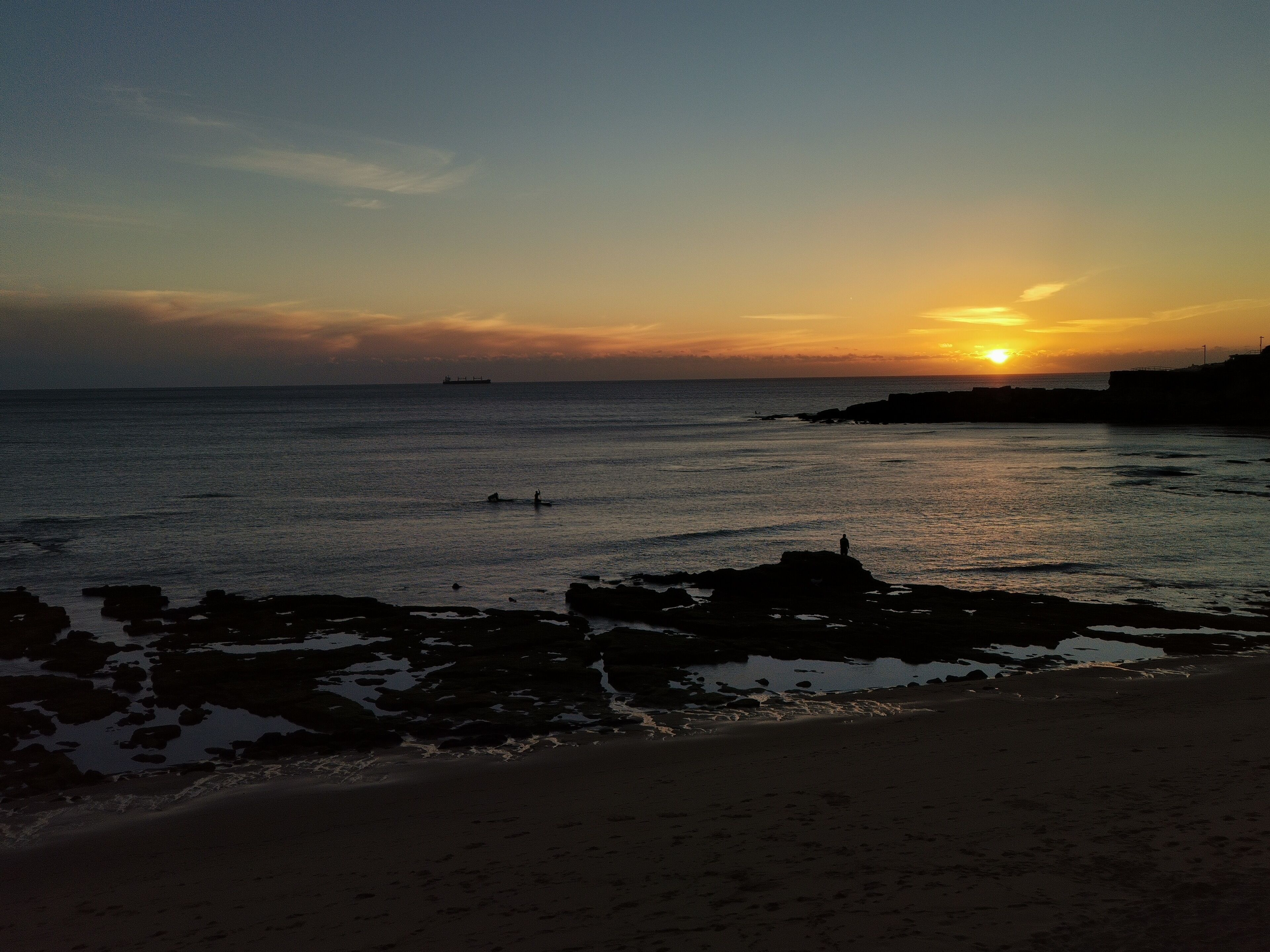A beach at sunset with calm waters, a distant ship, and rocky outcrops.