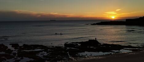 A beach at sunset with calm waters, a distant ship, and rocky outcrops.