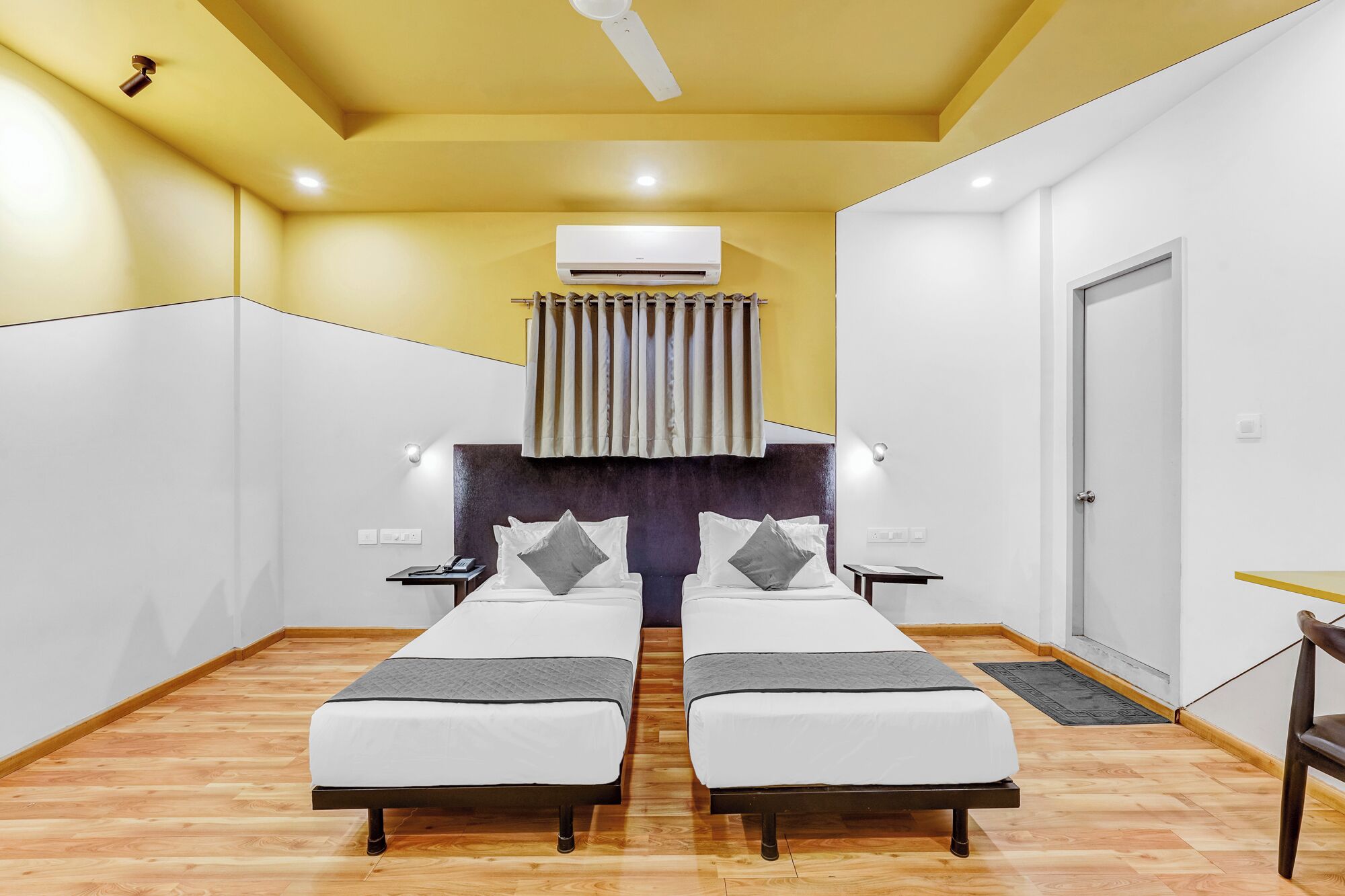 Una habitación con dos camas individuales, ventilador de techo, aire acondicionado, una mesita y una puerta.