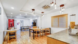 A modern hotel room with a wooden floor, a dining area with a table and chairs, a kitchen area with a coffee machine, and a large window with blinds.
