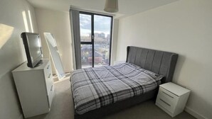 Un dormitorio con cama, televisor en una alacena, mesita de noche pequeña y ventana con vista a la ciudad.