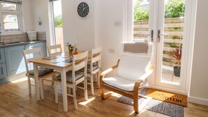 A bright, sunlit room with a wooden dining table, white chairs, a white armchair, and a clock on the wall. - Croeso (Holyhead)