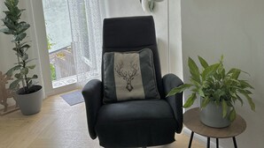 Un saló modern amb una butaca negra, una taula rodona petita amb una planta, un penjoll decoratiu i una vista per una porta de vidre.