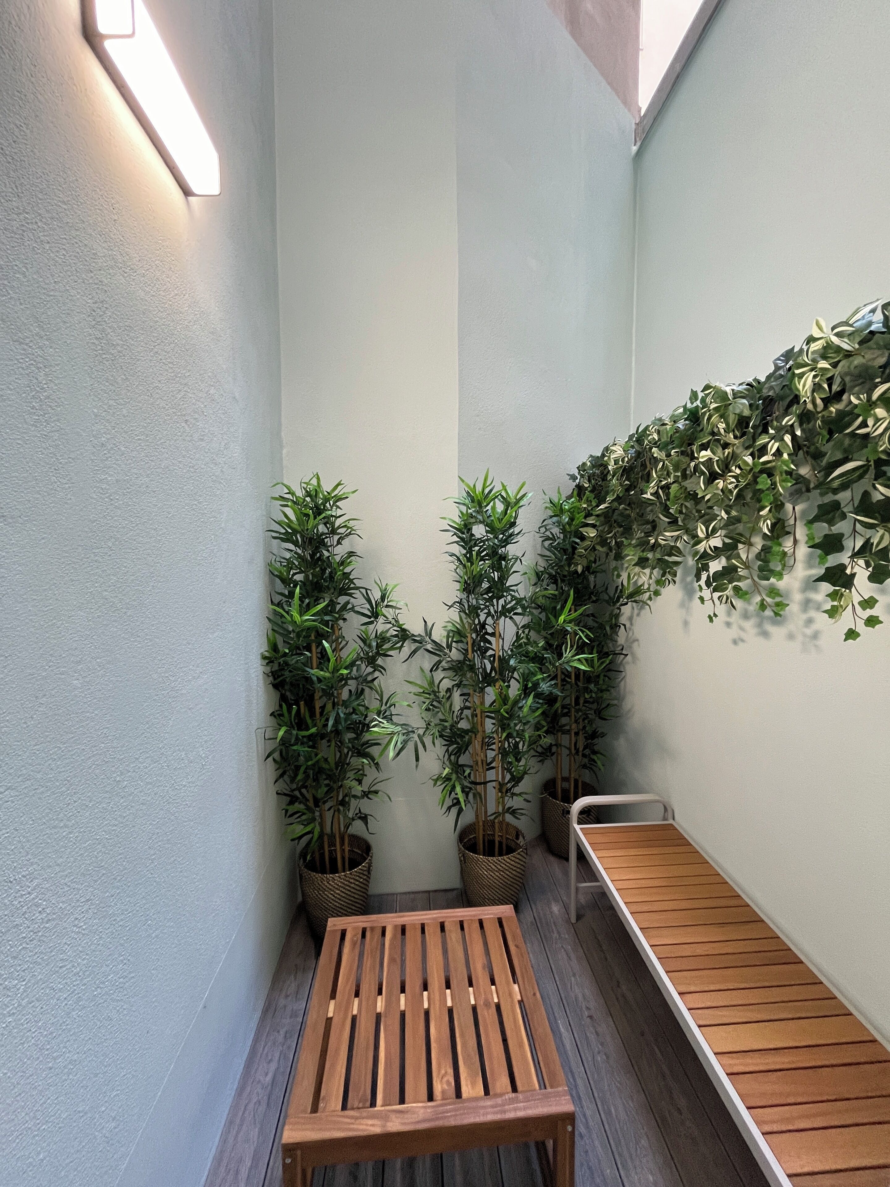 A small, enclosed outdoor space with a wooden bench, potted plants, and a wall-mounted light fixture.