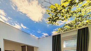 A compact hotel room with a bed, a small dining table, a kitchenette, and a view of a balcony with plants. - Street Living Hotel (Vienna)