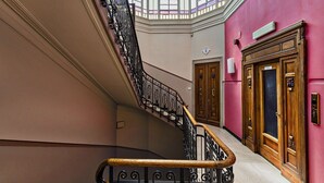 A grand staircase with ornate iron railings and wooden handrails, leading to a room with a large window and a red wall. - The Liberty Rooftop Terrace by Wonderful Italy (Genova)