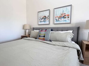 Un lit bien fait, avec une couette de couleur claire, une tête de lit grise et deux tableaux encadrés au-dessus.