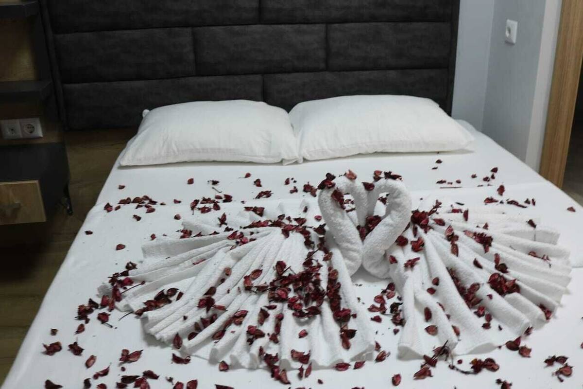 A neatly made bed with white linens and a towel arranged in a fan shape, surrounded by scattered rose petals.