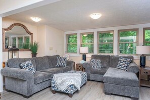 A living room with two grey sofas, a matching ottoman, a large mirror, and a window with a view of greenery.