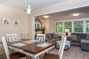 A dining area with a wooden table and chairs, a gray sofa, and a window with a view of trees.