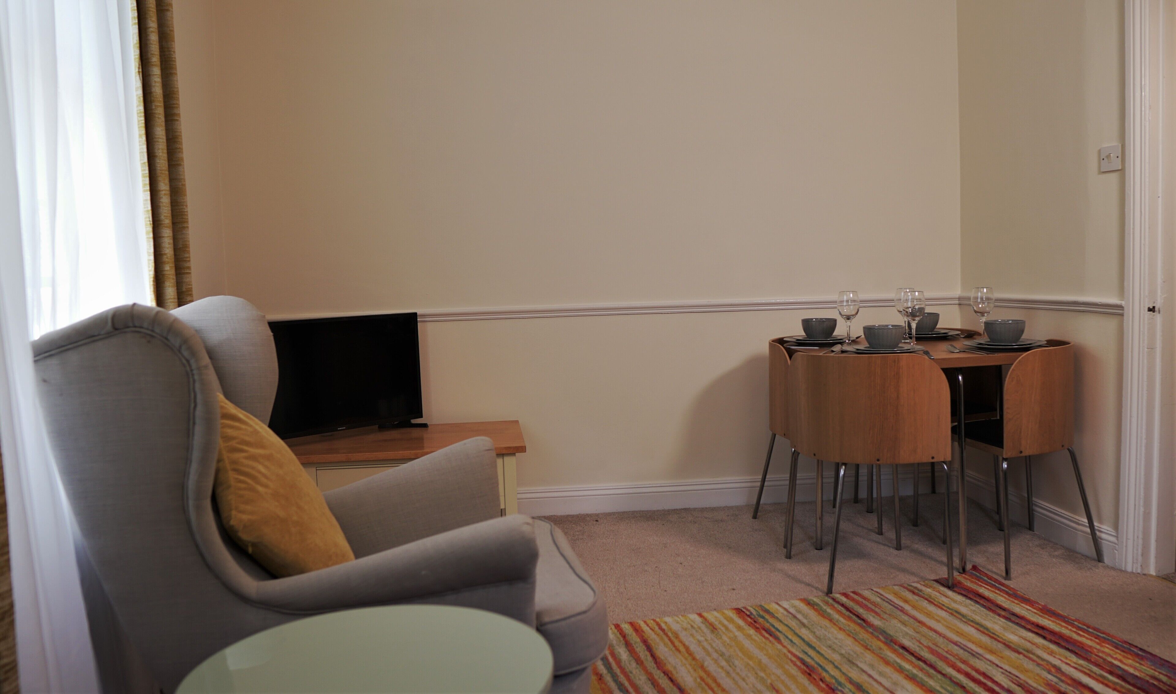 Una habitación pequeña y acogedora con un sillón gris, una mesa de madera para dos personas y un televisor sobre un mueble de madera.