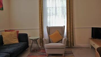 Una sala de estar con un sillón gris, un sofá negro, una mesita redonda y un televisor sobre un mueble de madera.