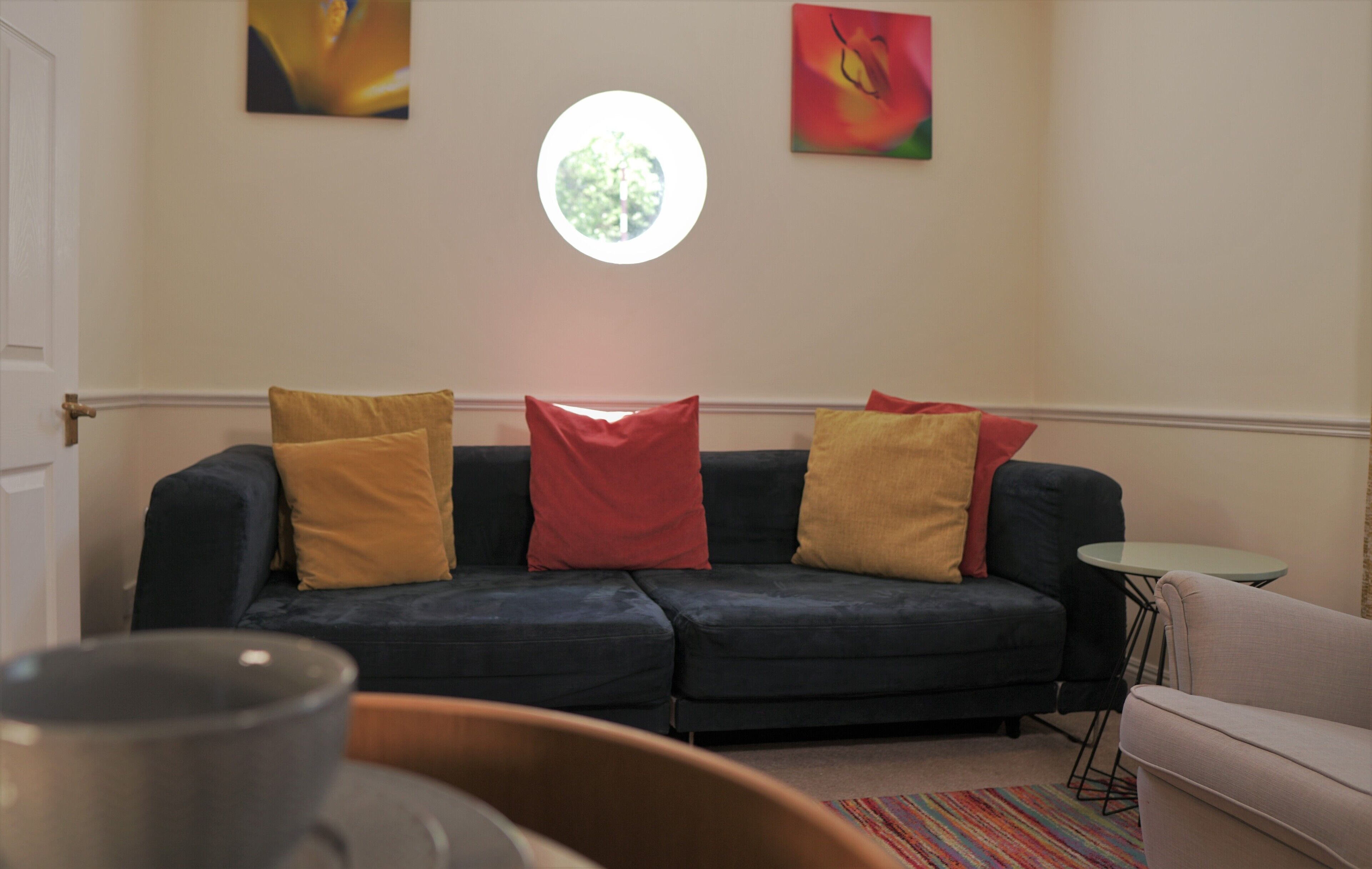 Un salon avec un canapé bleu foncé, des coussins rouges et jaunes, une fenêtre ronde et une petite table ronde.
