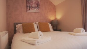 A neatly made bed with white linens and two brown pillows, a bedside lamp, and a painting of a snowy landscape on the wall. - Meadow Terrace (Shrewsbury)