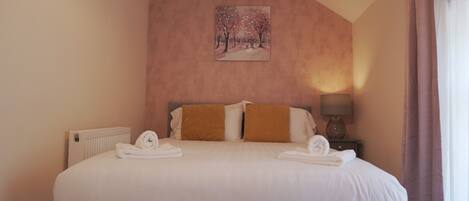 A neatly made bed with white and brown bedding, a radiator, a painting of trees, and a lamp on a bedside table.