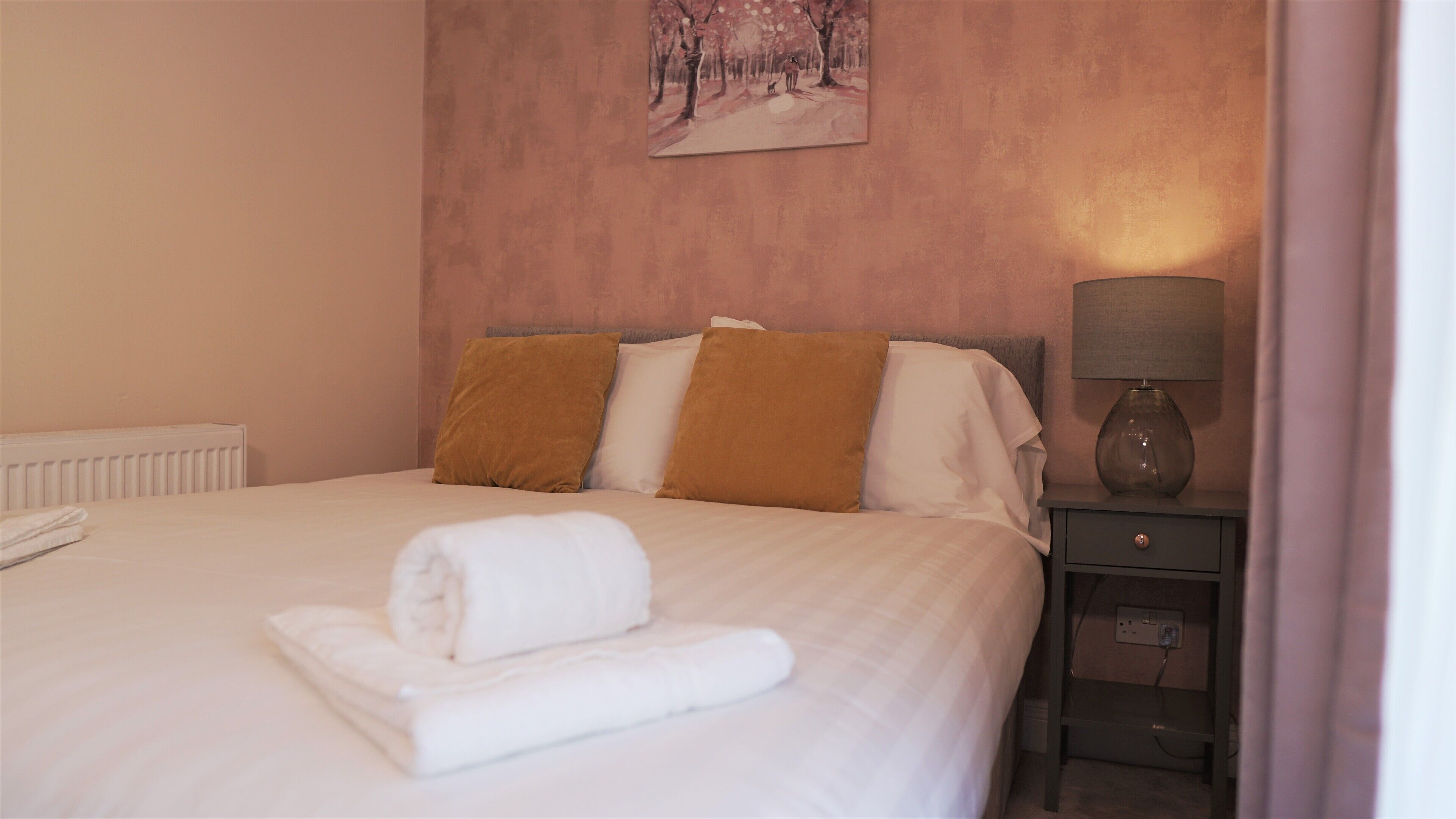 Una cama bien hecha con sábanas blancas, dos almohadas marrones y una toalla blanca enrollada sobre la cama. En la pared sobre la cama, hay un cuadro de un paisaje boscoso. Una lámpara está sobre una mesita de noche a la derecha.