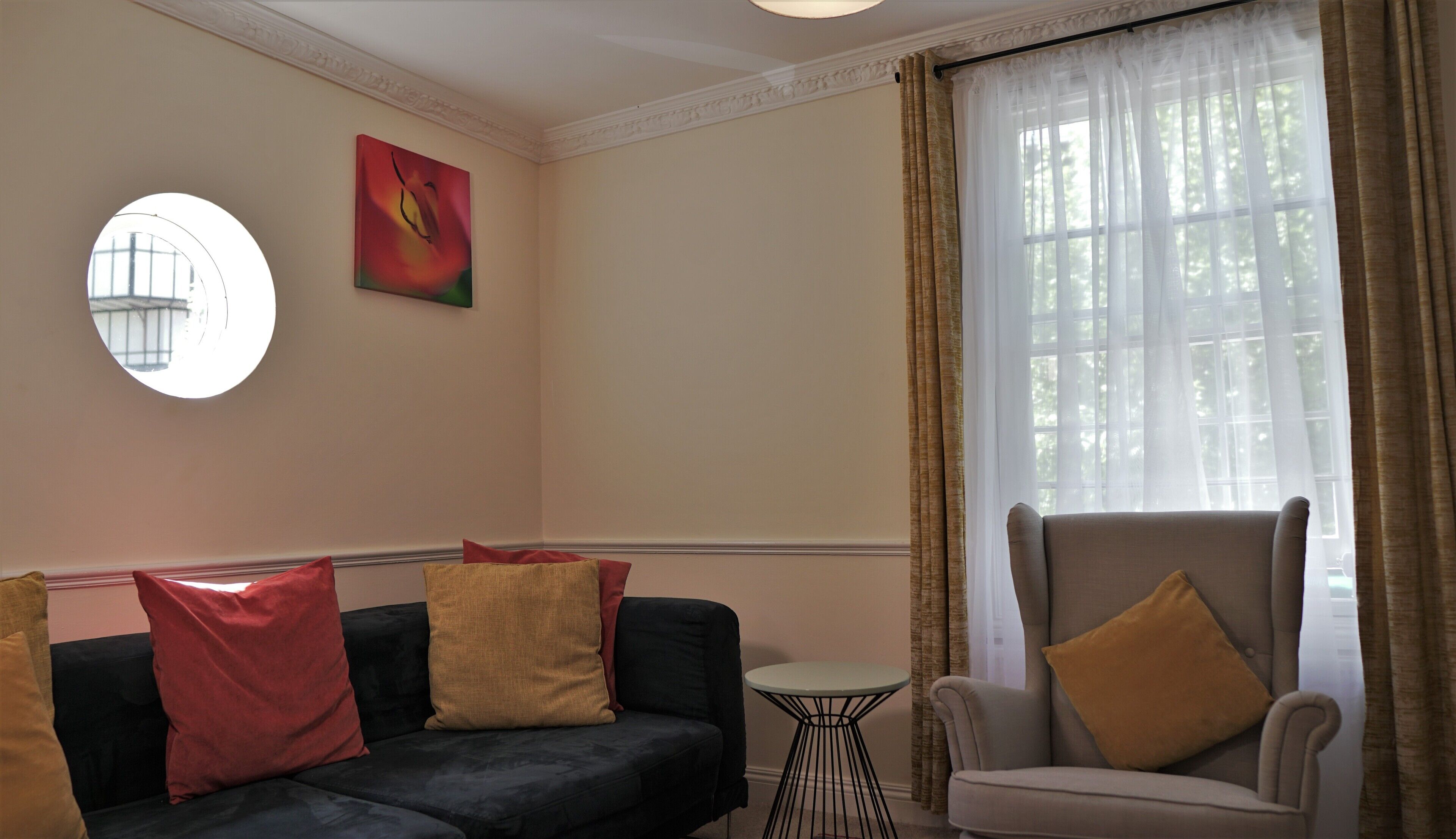Un salon avec un canapé foncé, un fauteuil beige et une petite table ronde. Il y a une fenêtre avec des rideaux et un tableau accroché au mur.