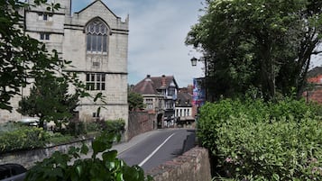A historic stone building with a prominent arched window, adjacent to a road lined with traditional houses and lush greenery.
