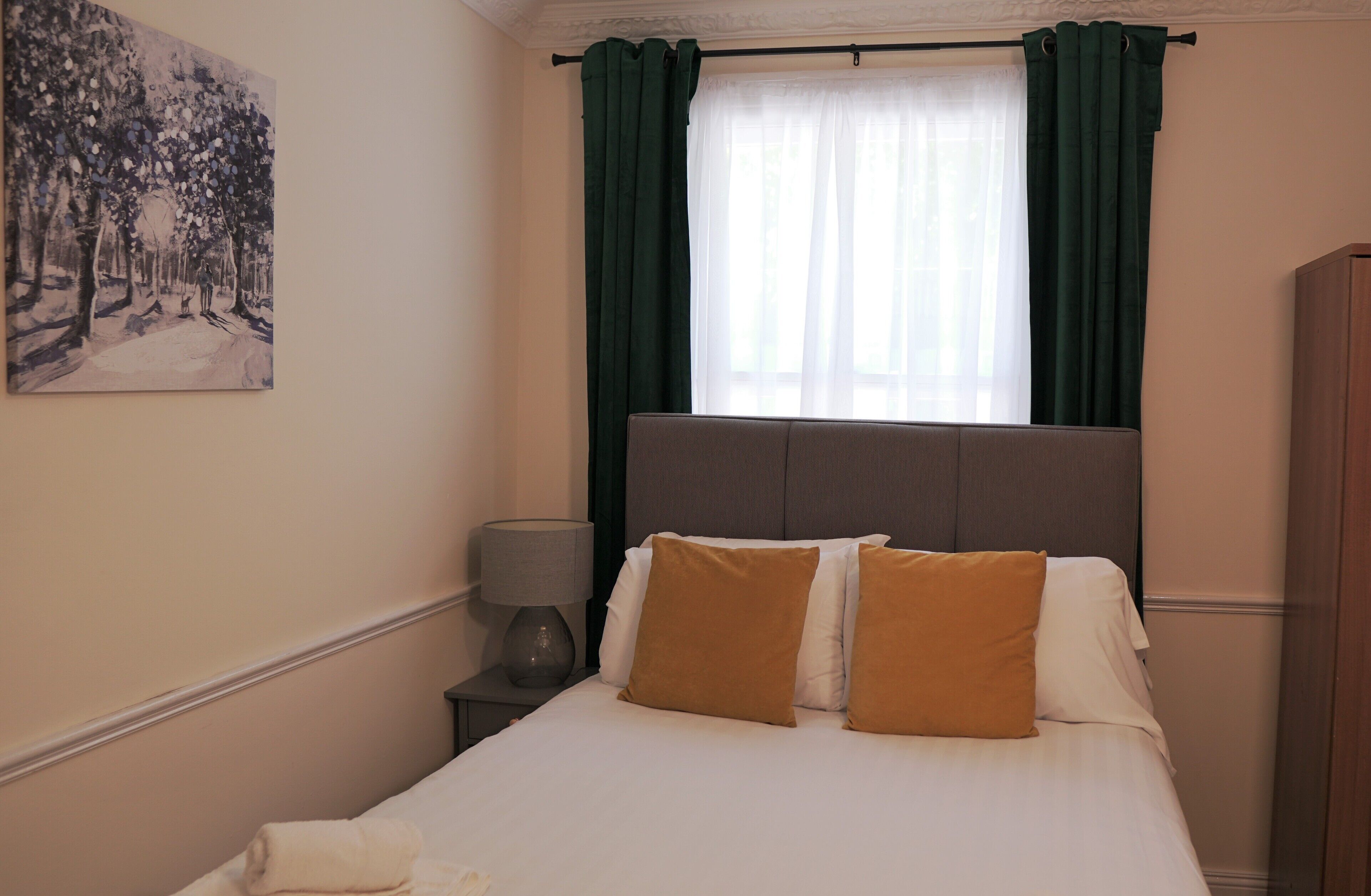 A bedroom with a bed, a nightstand, a lamp, a painting on the wall, and a window with curtains.