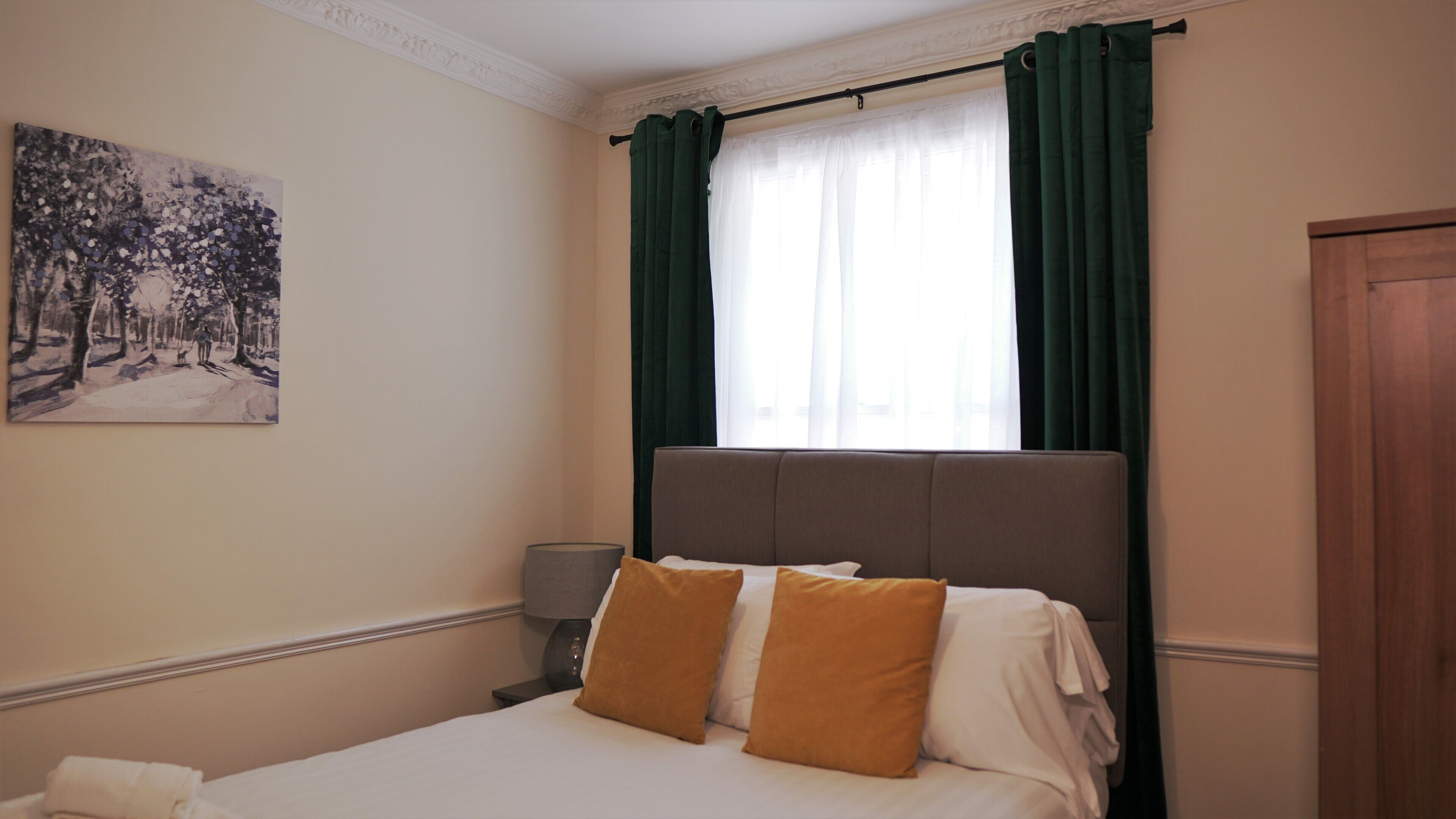 Habitación de hotel con cama, mesita de noche, lámpara, un cuadro y ventana con cortinas.