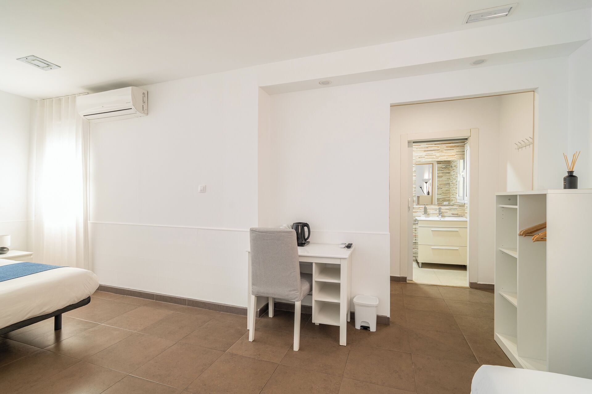 A hotel room with a bed, a desk, a chair, a small stool, a wardrobe, and a bathroom visible through an open door.