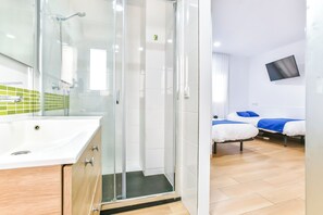 A modern bathroom with a glass shower enclosure, a white sink, and a green-tiled backsplash. There are two beds with blue bedding in the adjacent room.