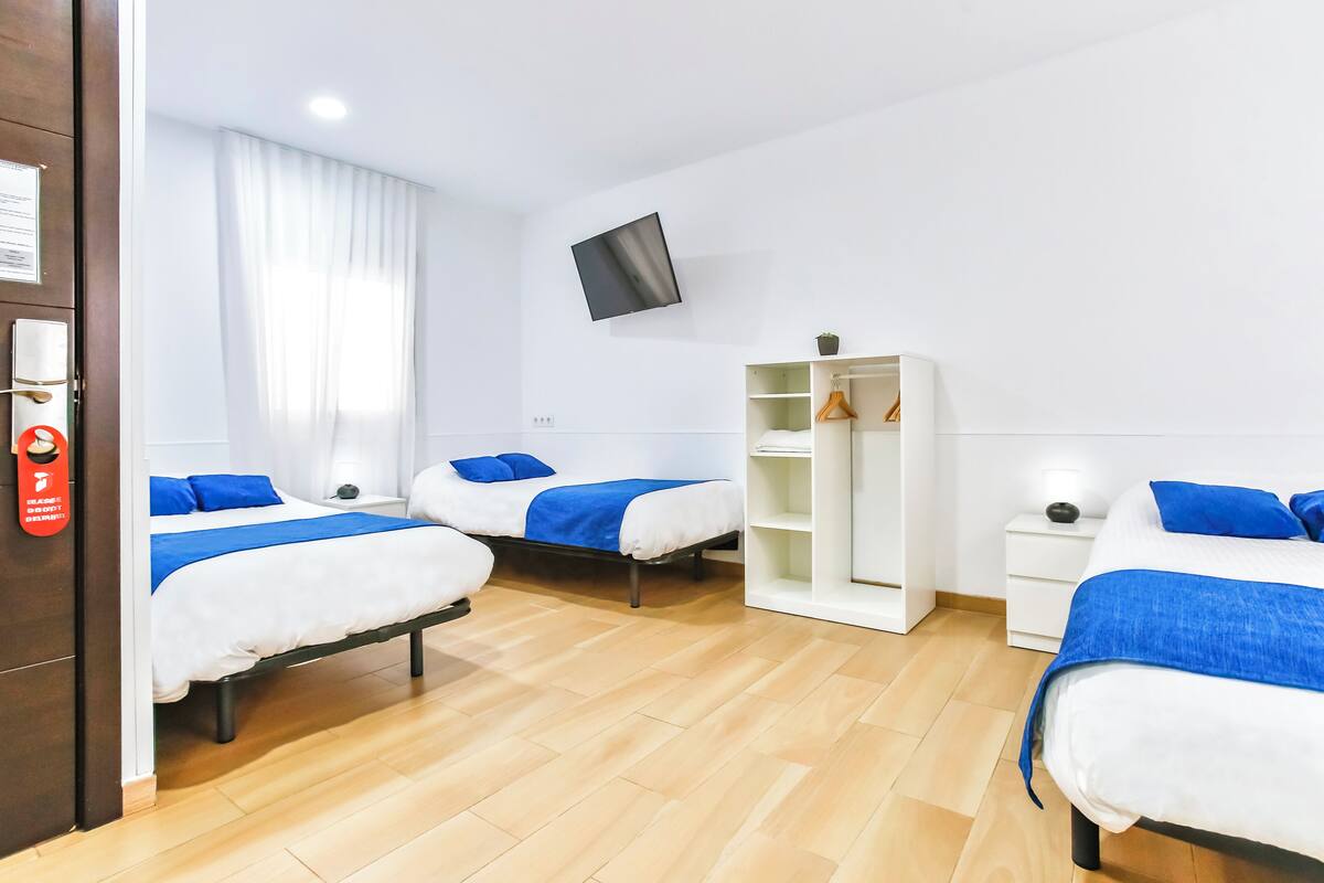 Habitación de hotel con tres camas, un televisor montado en la pared y un armario con estantes.