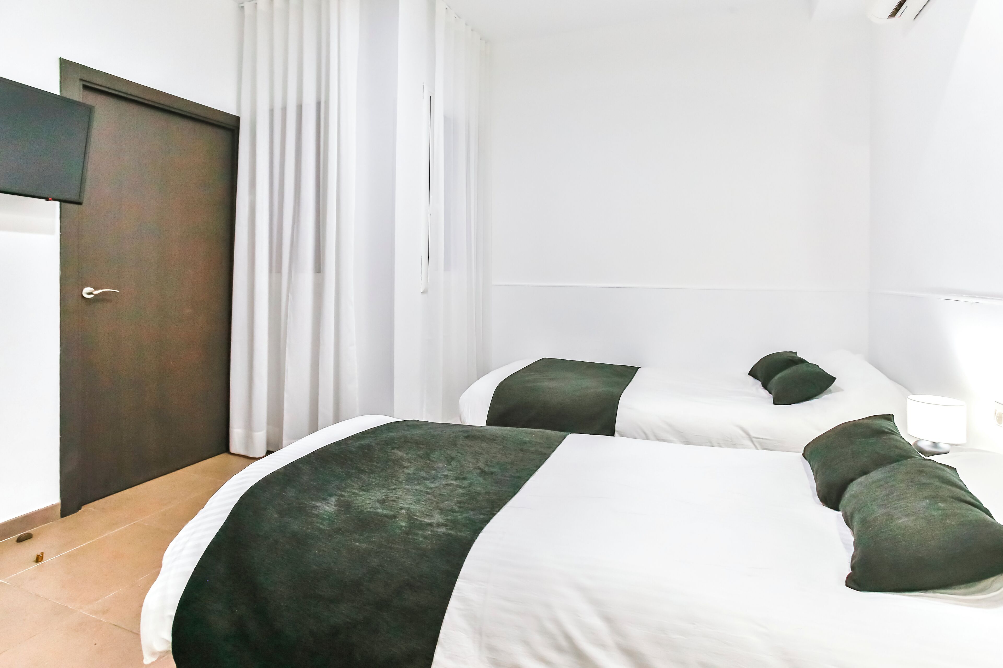 Habitación de hotel con dos camas, una puerta de madera oscura, cortinas blancas y una pared blanca.