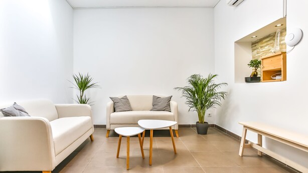 Una sala de estar moderna con un sofá color beige, dos mesas blancas, un banco de madera y plantas en macetas.