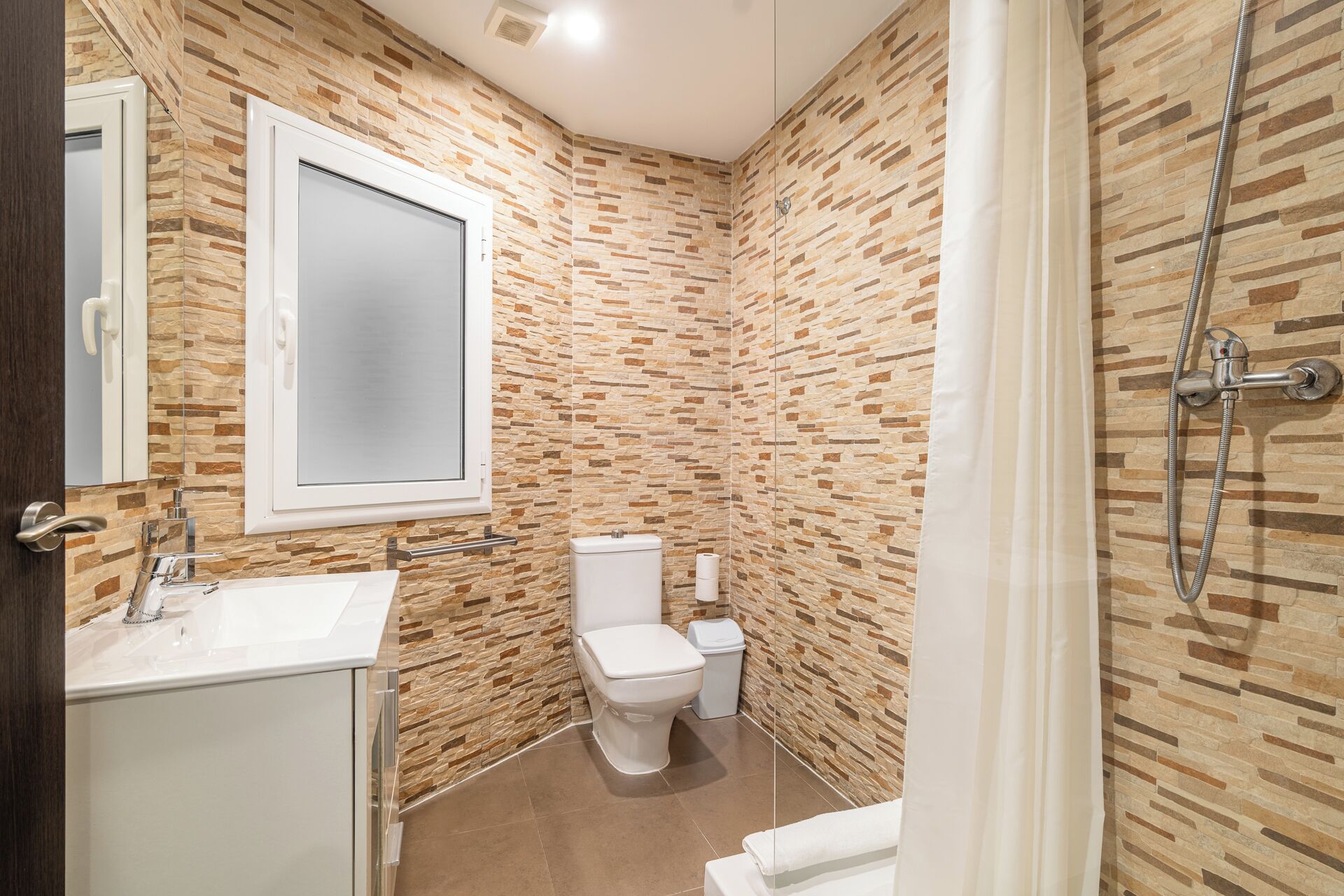 Une salle de bain avec un mur décoré d’un motif imitant la pierre, un lavabo blanc, des toilettes et une douche.