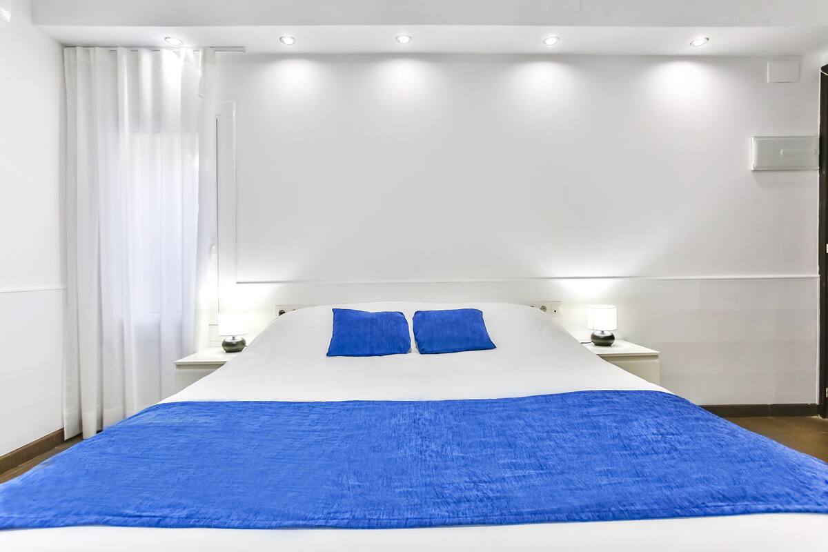 Habitación de hotel con una cama grande, paredes blancas y una colcha azul. Hay dos mesitas de noche con lámparas.