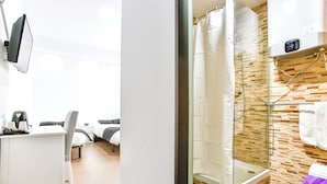Habitación de hotel con escritorio, silla, cama y zona de ducha con puerta de vidrio y pared de azulejos.