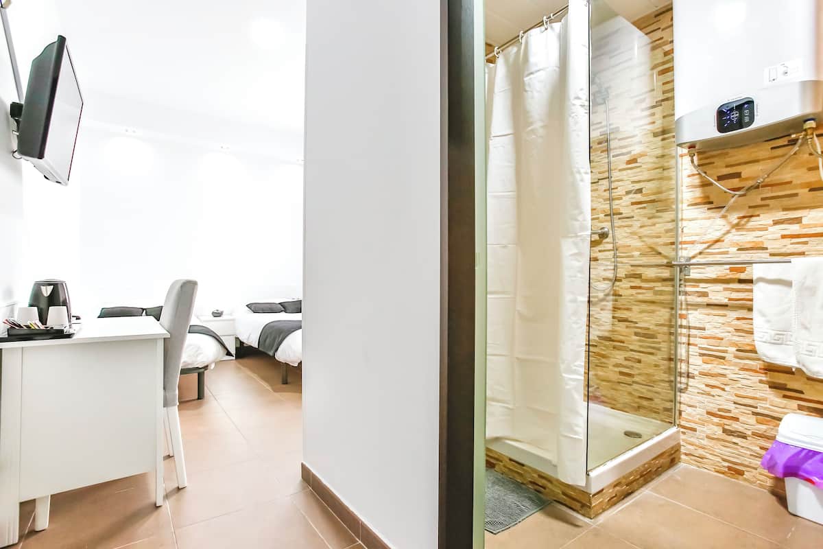 Habitación de hotel con escritorio, silla, cama y zona de ducha con puerta de vidrio y pared de azulejos.