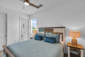 Un dormitorio con cama, mesita de noche, lámpara, ventilador de techo y ventana con persianas.