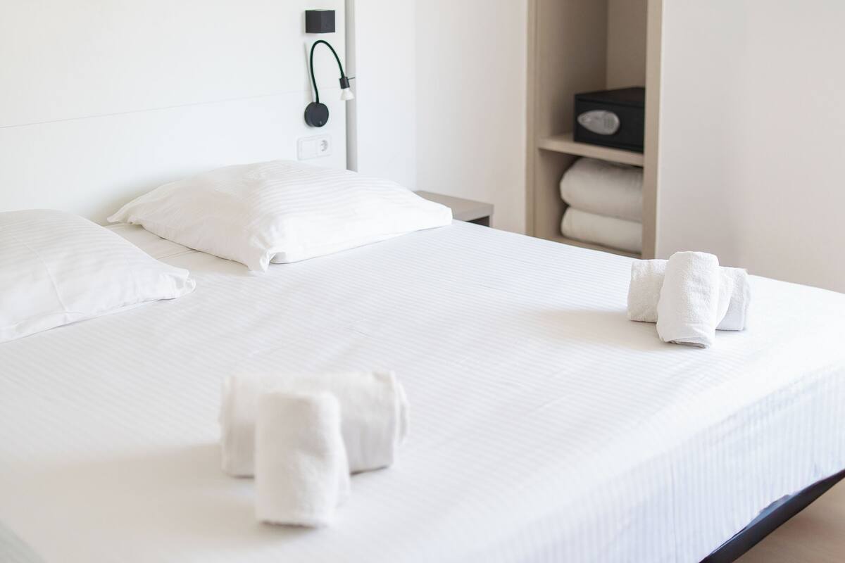 Una cama bien tendida con sábanas blancas y dos toallas sobre ella. Se observa un mesita de noche de madera con una lámpara y un enchufe. Al fondo, un armario de madera.