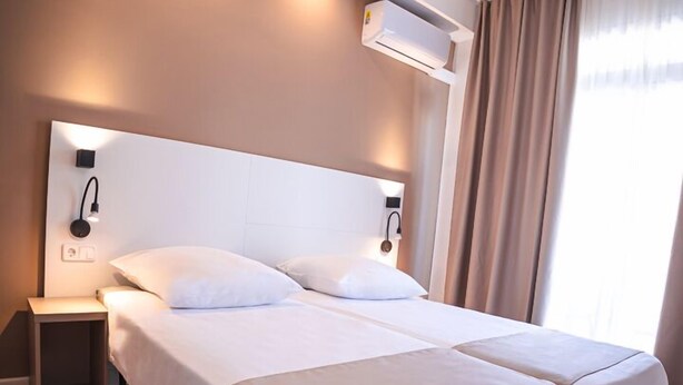 Una habitación de hotel con una cama grande, mesitas de noche, un equipo de aire acondicionado y una ventana con cortinas.