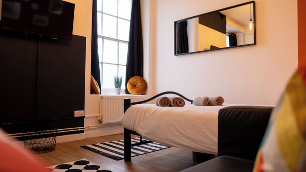 Un dormitorio con una cama, un armario negro, una ventana con cortinas, un radiador, un espejo y una planta en maceta.