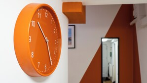 Un reloj naranja montado en la pared, con números y manecillas blancas, sobre un fondo blanco con diseño geométrico naranja y blanco.