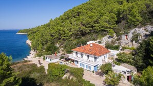 A coastal house with a red-tiled roof, surrounded by dense green trees and a clear blue sea.