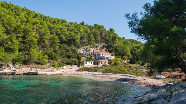 A coastal house with a red roof, surrounded by lush greenery and clear turquoise waters.
