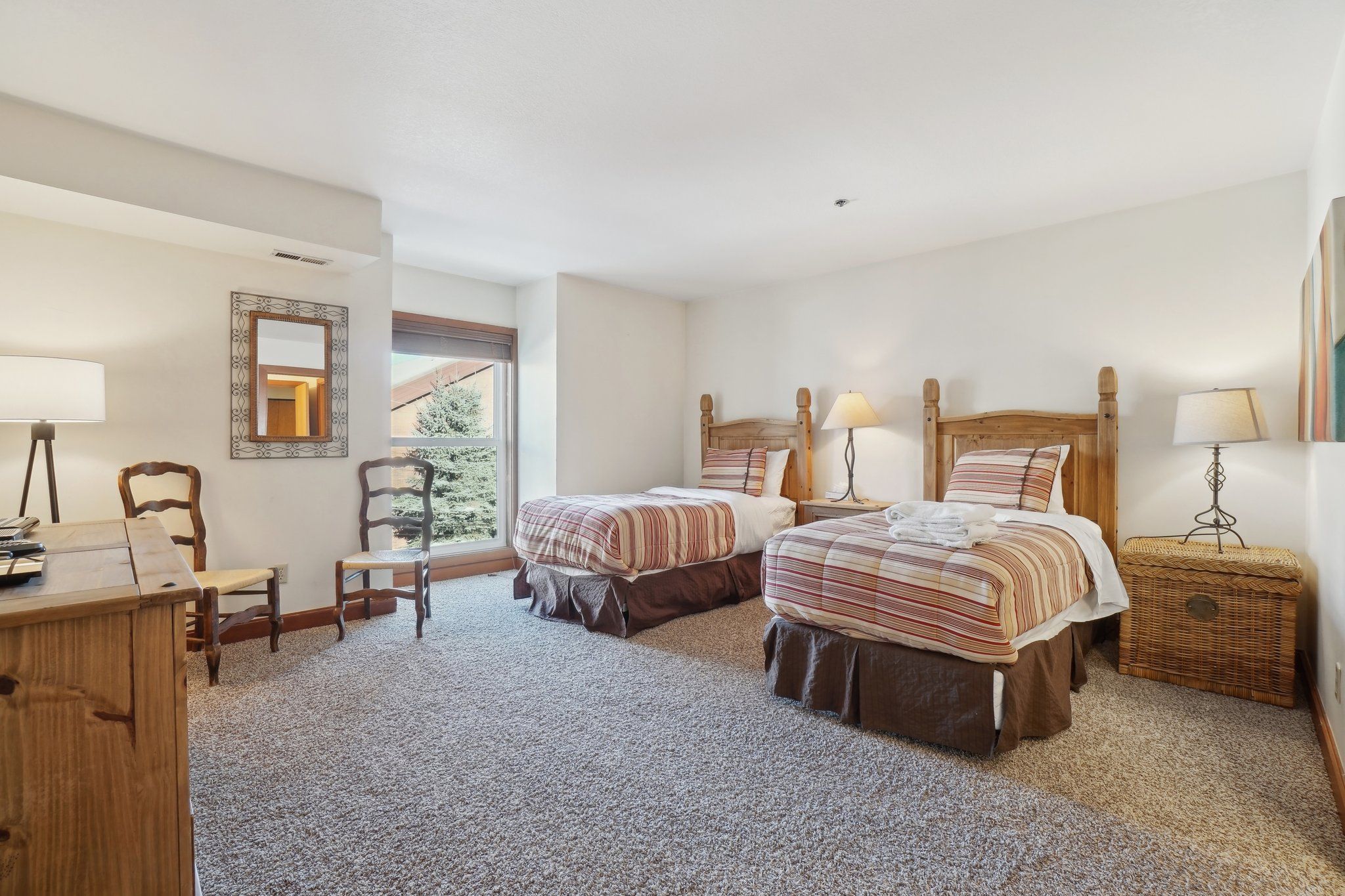 A bedroom with two beds, a desk, a chair, a mirror, and a window with a view of trees.