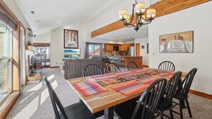 A spacious dining area with a wooden table and chairs, a kitchen in the background, and a large window providing natural light. - Silver Cliff 2BD - 2BA, Steps to PC Skiing and Old Town (Park City)