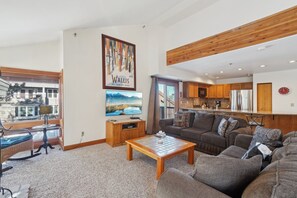 A modern living room with a sofa, a coffee table, and a television. There is a kitchen area with wooden cabinets and a refrigerator.