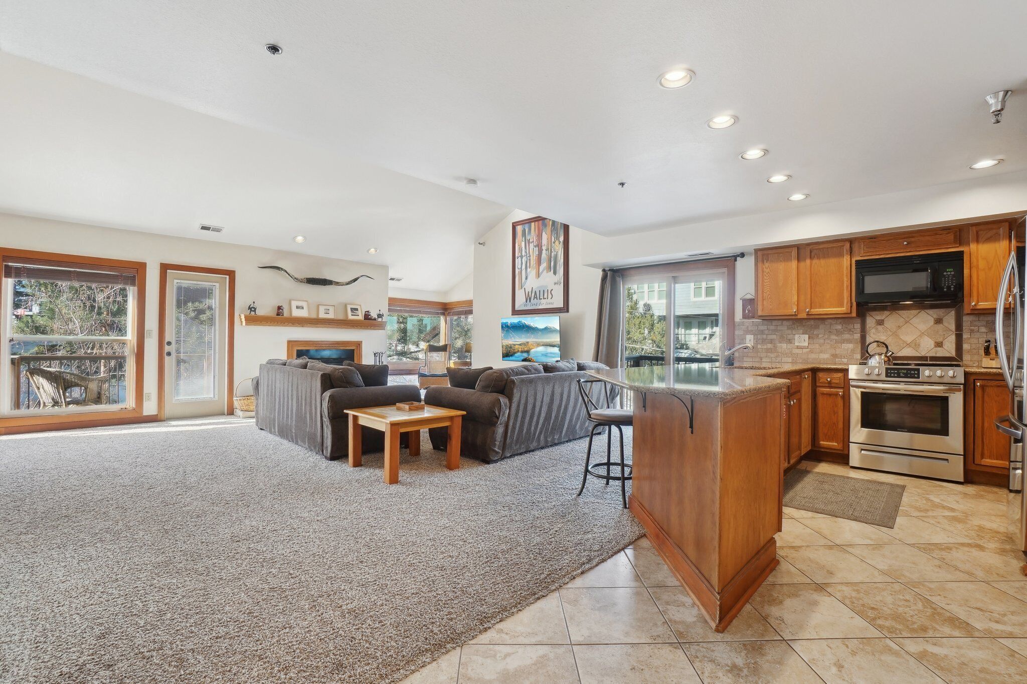 A spacious living area with a large gray carpet, a sofa, a coffee table, and a kitchen with wooden cabinets, a refrigerator, and a microwave.