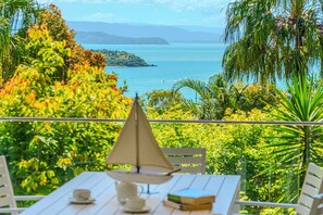 A table with a sail-shaped lamp, books, and a cup, overlooking a scenic view of a body of water and greenery. - Oasis 13, Oasis 13 on Hamilton Island (Hamilton Island)