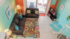 A living room with two dark sofas, a coffee table, and a colorful rug. There is a TV stand with a TV and a small table with a lamp. The room has teal walls and a window with a view of the outside.